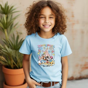Op de afbeelding: Een lichtblauw T-shirt met een kleurrijke afbeelding van Disney-personages voor een kasteel met vuurwerk. De tekst "Disney World" en "Est. 1971" staan ook op het shirt.