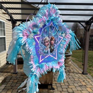 May include: A star-shaped pi&ntilde;ata with blue, pink, and white tissue paper fringe. The center features a Frozen-themed image of Elsa and Anna, with the word "FROZEN" in white. Decorative snowflakes are also present.