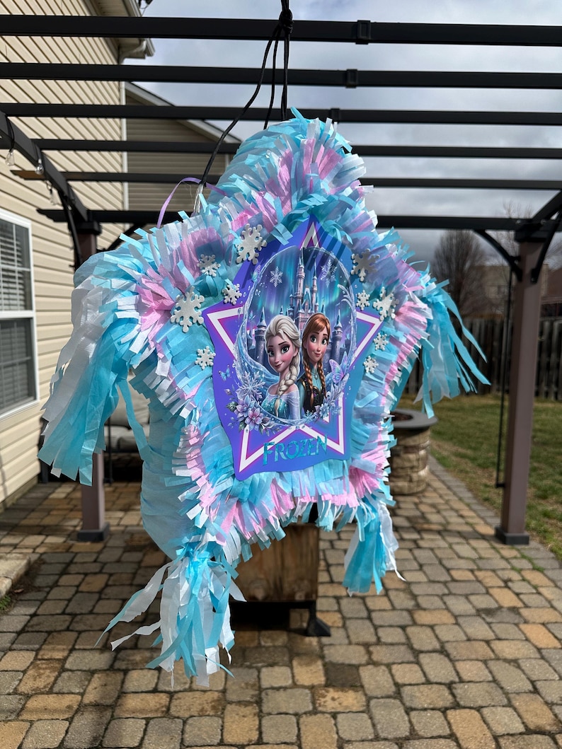 May include: A star-shaped pi&ntilde;ata with blue, pink, and white tissue paper fringe. The center features a Frozen-themed image of Elsa and Anna, with the word "FROZEN" in white. Decorative snowflakes are also present.