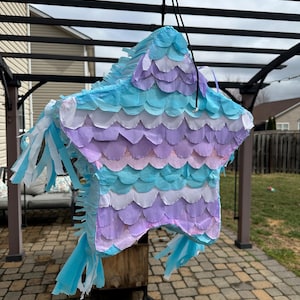 May include: A star-shaped pi&ntilde;ata in shades of blue, purple, and white, hanging from a black string. The pi&ntilde;ata is decorated with layered, scalloped paper and fringed streamers. It is suspended outdoors, suggesting a party or celebration.