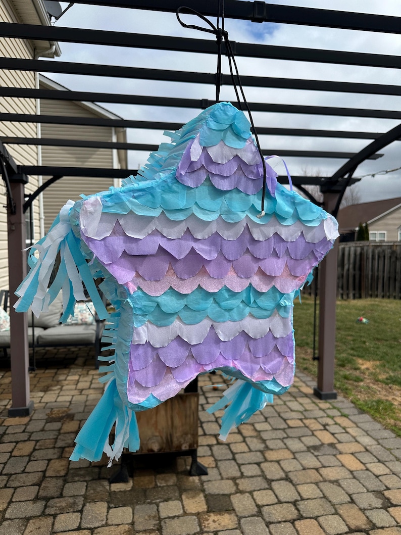 May include: A star-shaped pi&ntilde;ata in shades of blue, purple, and white, hanging from a black string. The pi&ntilde;ata is decorated with layered, scalloped paper and fringed streamers. It is suspended outdoors, suggesting a party or celebration.