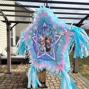 May include: A star-shaped pi&ntilde;ata with blue, pink, and white fringe. The center features a Frozen-themed image of Elsa and Anna in front of a castle. The pi&ntilde;ata is hanging from a black string, ready for a party.