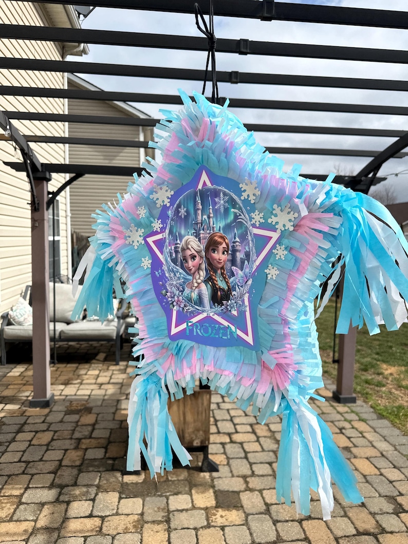 May include: A star-shaped pi&ntilde;ata with blue, pink, and white fringe. The center features a Frozen-themed image of Elsa and Anna in front of a castle. The pi&ntilde;ata is hanging from a black string, ready for a party.
