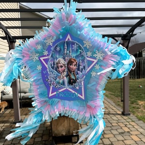 May include: A star-shaped pi&ntilde;ata with blue, pink, and white fringe. The center features a Frozen-themed image of Elsa and Anna in front of a castle. The word "FROZEN" is printed on the pi&ntilde;ata.
