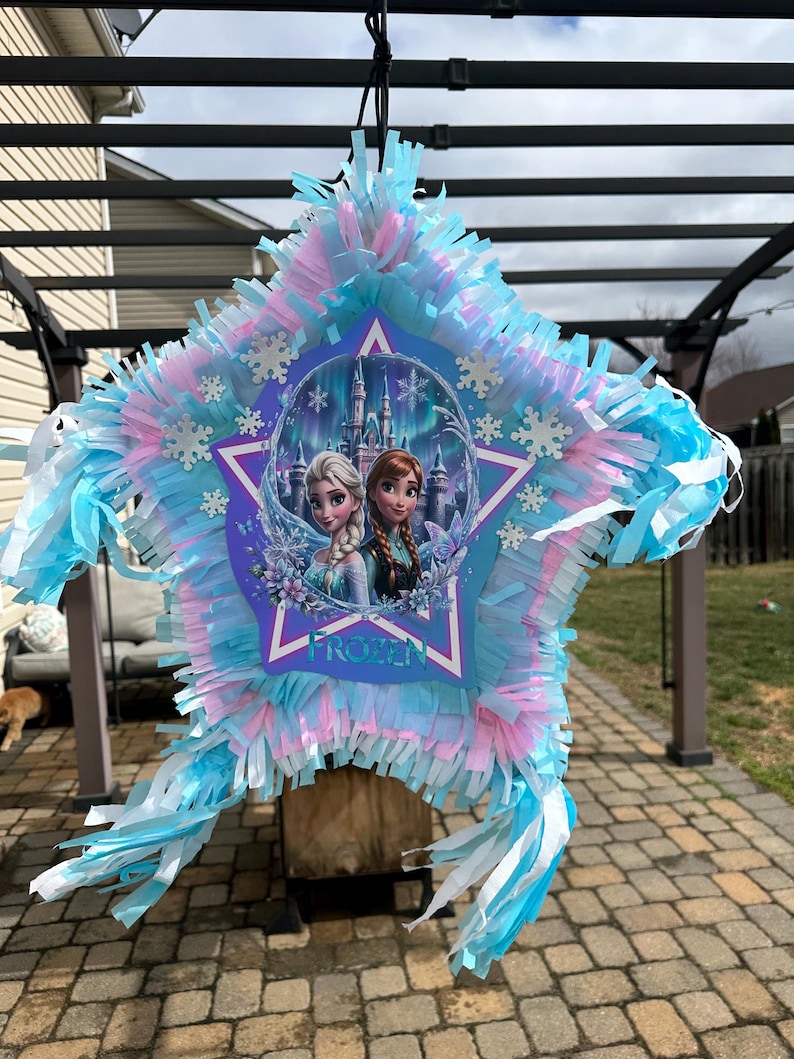 May include: A star-shaped pi&ntilde;ata with blue, pink, and white fringe. The center features a Frozen-themed image of Elsa and Anna in front of a castle. The word "FROZEN" is printed on the pi&ntilde;ata.