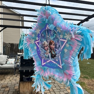 May include: A star-shaped pi&ntilde;ata with blue, pink, and white tissue paper fringe. The center features a Frozen-themed image of Elsa and Anna, with the word "FROZEN" below. The pi&ntilde;ata is hanging outdoors.