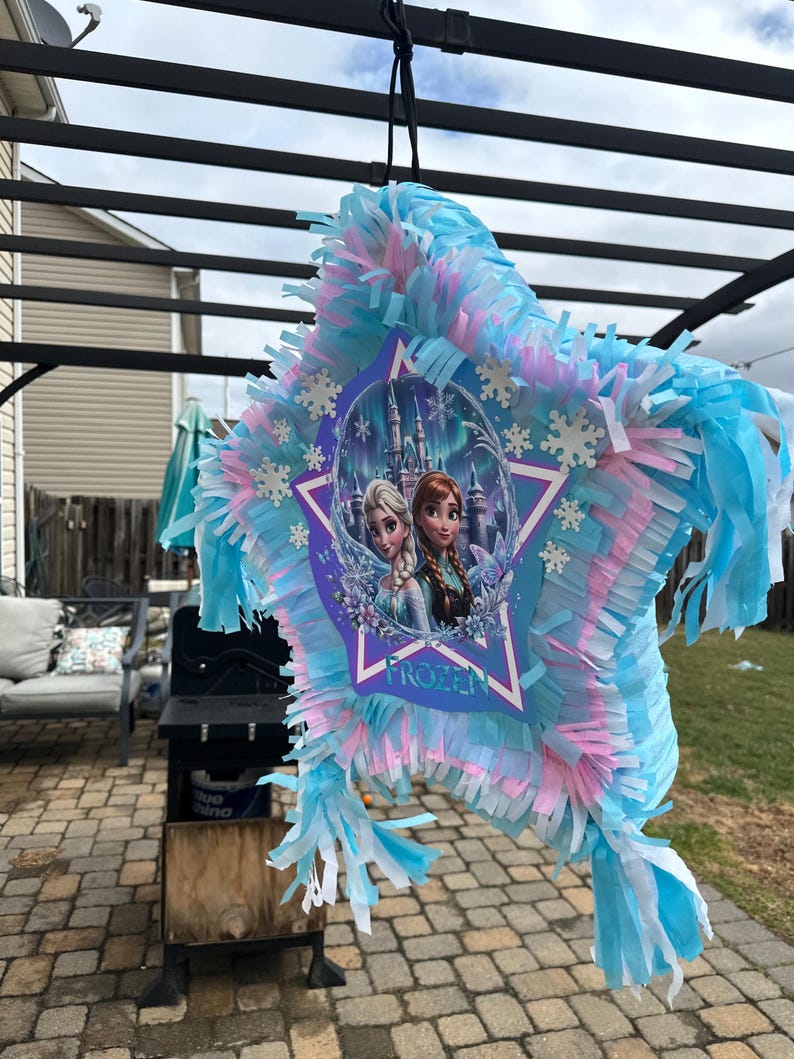 May include: A star-shaped pi&ntilde;ata with blue, pink, and white tissue paper fringe. The center features a Frozen-themed image of Elsa and Anna, with the word "FROZEN" below. The pi&ntilde;ata is hanging outdoors.
