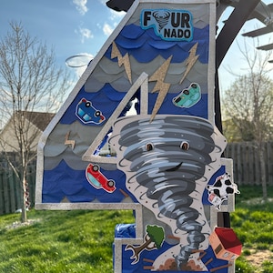 May include: A large, number four decoration with a "FOUR-NADO" theme. The number is blue and silver with a cartoon tornado, cars, lightning bolts, a house, a tree, and a cow. The decoration is outdoors in front of green grass.
