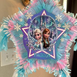 May include: A star-shaped pi&ntilde;ata featuring characters from the movie "Frozen." The pi&ntilde;ata is decorated with blue, pink, and white fringed paper. The center image shows Elsa and Anna in front of a castle, with the word "FROZEN" below.