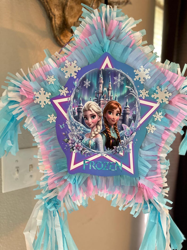 May include: A star-shaped pi&ntilde;ata featuring characters from the movie "Frozen." The pi&ntilde;ata is decorated with blue, pink, and white fringed paper. The center image shows Elsa and Anna in front of a castle, with the word "FROZEN" below.