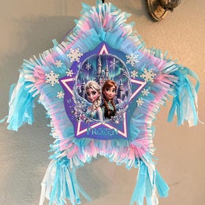 May include: Star-shaped pi&ntilde;ata with blue, pink, and white fringe. The center features a purple star with the word "FROZEN" and images of Elsa and Anna from the Disney movie. White snowflake accents are scattered around the star.