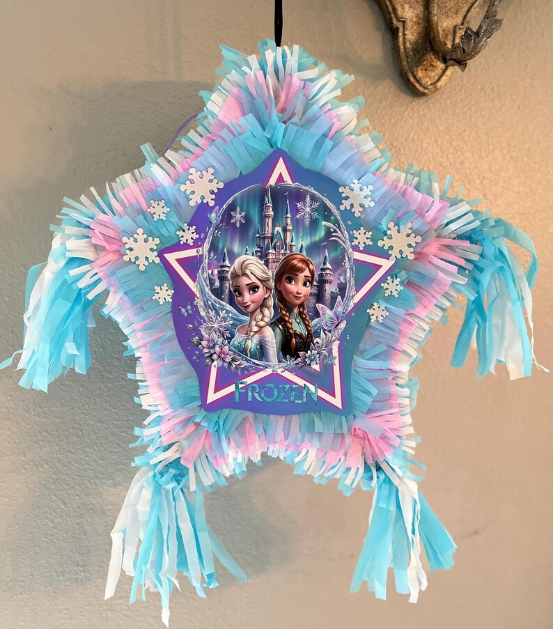 May include: Star-shaped pi&ntilde;ata with blue, pink, and white fringe. The center features a purple star with the word "FROZEN" and images of Elsa and Anna from the Disney movie. White snowflake accents are scattered around the star.
