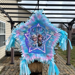 May include: A star-shaped pi&ntilde;ata with blue, pink, and white fringe. The center features a Frozen-themed design with Elsa and Anna, snowflakes, and the word "Frozen". The pi&ntilde;ata is suspended from a black structure.
