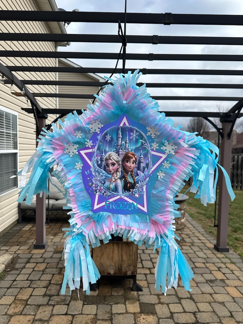 May include: A star-shaped pi&ntilde;ata with blue, pink, and white fringe. The center features a Frozen-themed design with Elsa and Anna, snowflakes, and the word "Frozen". The pi&ntilde;ata is suspended from a black structure.
