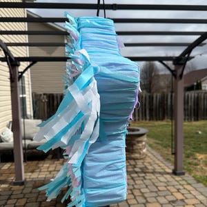 May include: A light blue pi&ntilde;ata with layered crepe paper construction. The pi&ntilde;ata is decorated with white, light blue, and pink fringe. It is suspended from a black cord, ready for a party or celebration.