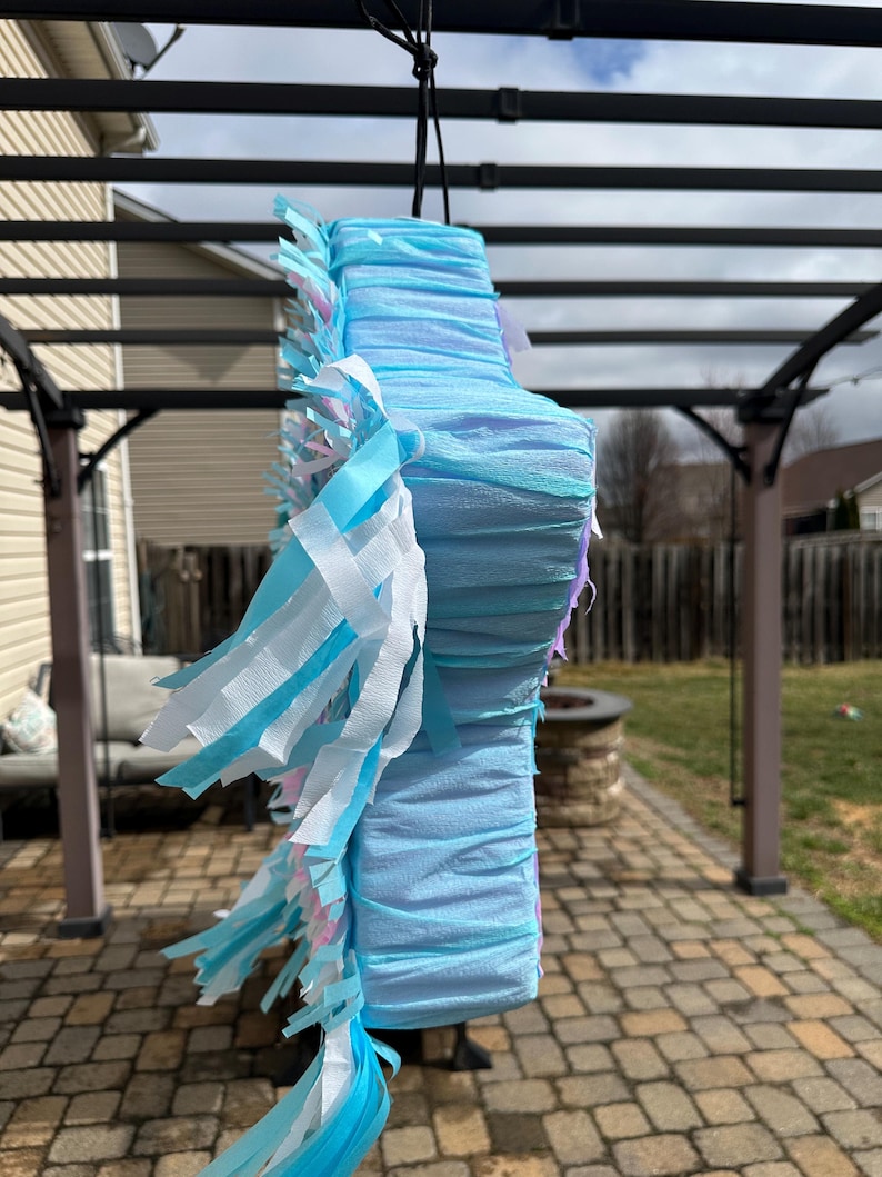 May include: A light blue pi&ntilde;ata with layered crepe paper construction. The pi&ntilde;ata is decorated with white, light blue, and pink fringe. It is suspended from a black cord, ready for a party or celebration.