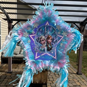 May include: Star-shaped pi&ntilde;ata with a Frozen theme. The pi&ntilde;ata features Elsa and Anna, surrounded by a star-shaped frame. The pi&ntilde;ata is decorated with blue, pink, and white streamers and snowflake accents. It is hanging outdoors.
