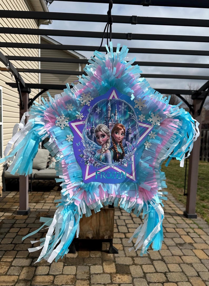 May include: Star-shaped pi&ntilde;ata with a Frozen theme. The pi&ntilde;ata features Elsa and Anna, surrounded by a star-shaped frame. The pi&ntilde;ata is decorated with blue, pink, and white streamers and snowflake accents. It is hanging outdoors.