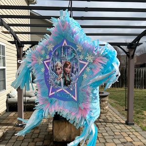 May include: A star-shaped pi&ntilde;ata with blue, pink, and white tissue paper fringe. The center features a graphic of two characters from the movie "Frozen" in front of a castle. The word "FROZEN" is printed on the pi&ntilde;ata.