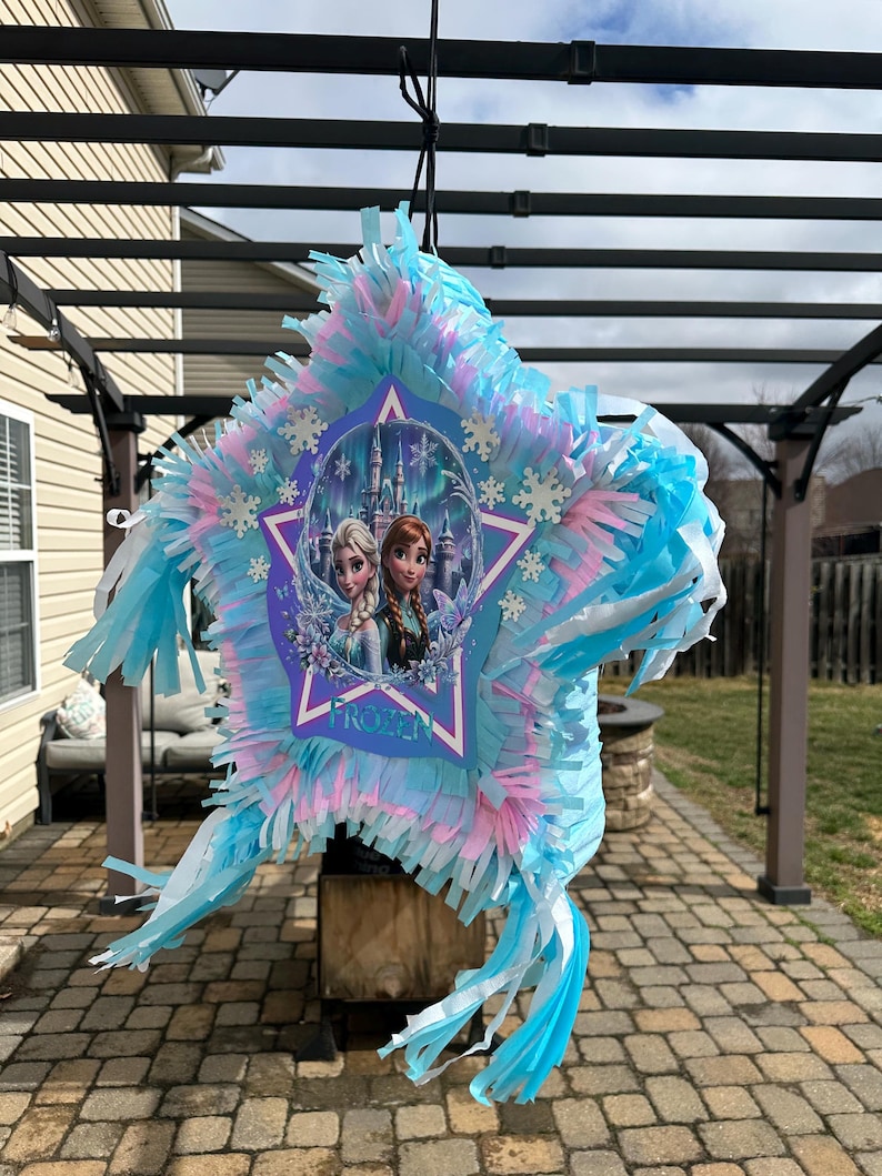 May include: A star-shaped pi&ntilde;ata with blue, pink, and white tissue paper fringe. The center features a graphic of two characters from the movie "Frozen" in front of a castle. The word "FROZEN" is printed on the pi&ntilde;ata.