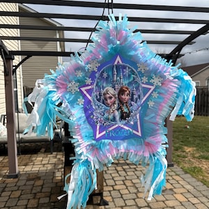 May include: A star-shaped pi&ntilde;ata with blue, pink, and white fringe. The center features an image of Elsa and Anna from the movie "Frozen" with the word "Frozen" printed below. Decorative snowflakes are attached.