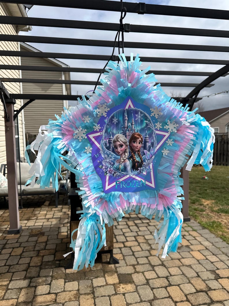 May include: A star-shaped pi&ntilde;ata with blue, pink, and white fringe. The center features an image of Elsa and Anna from the movie "Frozen" with the word "Frozen" printed below. Decorative snowflakes are attached.