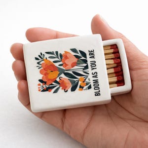 Peut inclure: Une boîte d'allumettes blanche avec un motif floral orange, rouge et vert, ouverte pour révéler les allumettes. Le texte "BLOOM AS YOU ARE" est imprimé verticalement sur le côté. Les allumettes ont des têtes rouges.