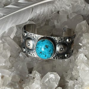 May include: A silver cuff bracelet with a textured surface and circular embellishments. The bracelet features a large, round turquoise stone in the centre. A white feather and crystals are in the background.