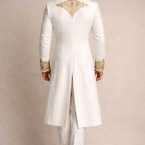May include: A white formal suit with gold embroidery. The long coat features ornate gold detailing on the shoulders and cuffs. The suit includes matching white trousers and a long coat that reaches the ankles. The back of the coat has a split.
