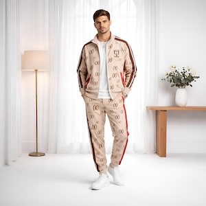 Op de afbeelding: Een beige trainingspak met een herhaald "00" patroon en bruine accenten. De jas heeft een witte kraag en bruine strepen op de mouwen. De broek heeft een rode streep aan de zijkant. De persoon draagt witte sneakers.