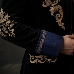 Puede incluir: Chaqueta de terciopelo negro con bordados dorados en las mangas y hombros. Los pu&ntilde;os son de color azul oscuro con un ribete dorado. El dise&ntilde;o ornamentado sugiere una prenda formal o teatral.