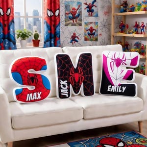 May include: Three letter-shaped pillows spelling "SME" on a white couch. The "S" is red and blue with "MAX" printed on it. The "M" is black with a red spider and "JACK" printed on it. The "E" is white and pink with "EMILY" printed on it.