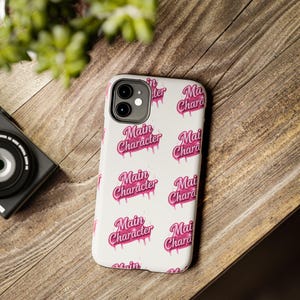 Puede incluir: Una funda de teléfono blanca con un gráfico rosa repetido que dice "Main Character" en una fuente goteante. La funda del teléfono está sobre una superficie de madera, con una cámara y vegetación en el fondo.