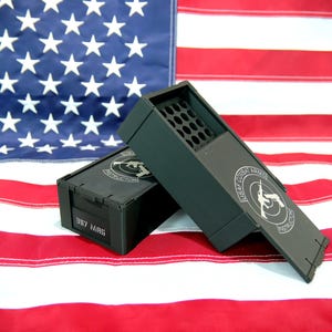 May include: Two matte black rectangular ammunition storage boxes with a grid of circular holes on the top. The boxes feature a circular logo with the text USAF COMBAT ARMS INSTRUCTOR and the side of one box is labeled with the text 357 MAG.