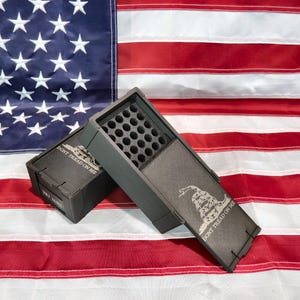 May include: Two dark gray rectangular ammunition storage boxes with sliding lids. Each box features a white graphic of a coiled rattlesnake and the text DONT TREAD ON ME. The open box reveals a grid of circular holes for holding individual cartridges.
