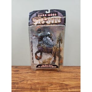 Peut inclure: Une figurine articulée centaure Spawn Dark Ages dans son emballage d'origine. La figurine présente un torse à la peau sombre avec une armure argentée, un corps de cheval avec du tissu violet et une longue lance. L'emballage porte le texte Spawn.