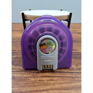 May include: A purple View-Master viewer featuring a Dora the Explorer image on the front. The viewer has a white rectangular button with the View-Master logo. The viewer is on a wooden surface.