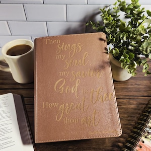 May include: A rose-colored Bible with gold lettering that reads "Then sings my soul, my savior God to Thee, How great thou art." The image includes a cup of coffee, a plant, and an open book.