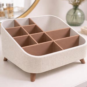 May include: A beige and brown organiser with nine compartments, suitable for storing cosmetics or small items. The organiser has a textured exterior, a curved design, and rests on small wooden legs. The interior compartments are a warm brown colour.