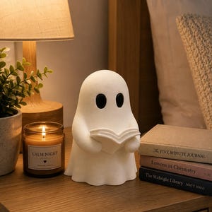 Peut inclure: Une lampe blanche en forme de fantôme avec des yeux ovales noirs, tenant un livre ouvert. La lampe est posée sur une surface en bois à côté d'une bougie allumée dans un pot en verre étiqueté CALM NIGHT et une pile de trois livres intitulés The FIVE MINUTE JOURNAL, Lessons in Chemistry et The Midnight Library.