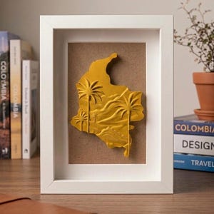 Peut inclure: Un cadre rectangulaire blanc contenant une carte en relief tridimensionnelle jaune de la Colombie. La carte présente des palmiers en relief et des détails de terrain montagneux sur un fond brun uni, encadrée par une bordure blanche moderne.