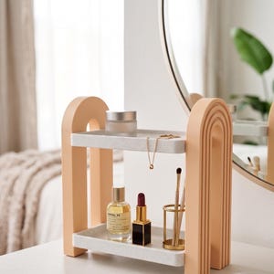 May include: A two-tiered peach-coloured arched shelf with white speckled rectangular trays. The top shelf holds a small frosted jar and a gold chain necklace. The bottom shelf contains a glass perfume bottle, a lipstick, and a gold brush holder.