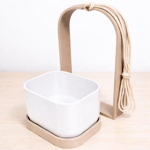 May include: A white square planter with a matching beige tray, alongside a beige arch-shaped hanger and natural rope for suspension. The minimalist design is suitable for indoor plants.