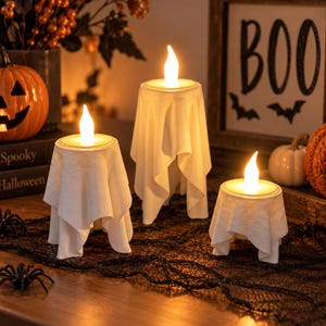 May include: Three LED candles of varying heights, each draped in a white fabric ghost costume. The candles emit a warm, glowing light from their flame-shaped tops. The ghost figures are positioned on a black lace runner, creating a festive display.