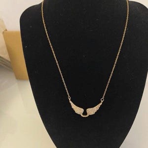 Peut inclure: Un collier doré avec une chaîne délicate et un pendentif en forme d'ailes d'ange. Les ailes sont ornées de petites pierres scintillantes, créant un effet chatoyant. Le collier est présenté sur un mannequin noir.