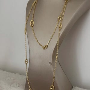 Peut inclure: Un collier doré avec un motif de chaîne et des maillons ovales. Le collier est présenté sur un mannequin. Le collier a un fermoir à bascule et est superposé.