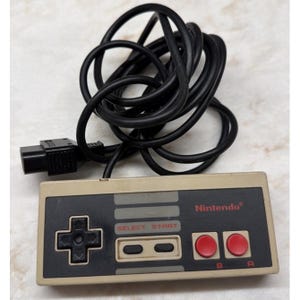 May include: A vintage Nintendo controller with a beige body, black directional pad, and red A and B buttons. The controller has a black cable and connector. The Nintendo logo is visible in red.