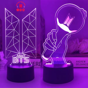 以下が含まれることがあります： 2つの紫色のLEDナイトライト。1つは「ARMY BTS」の文字が入った幾何学模様のデザインです。もう1つは、光を手に持っている様子を描いています。どちらも黒い台座にあります。ライトは本棚にあります。