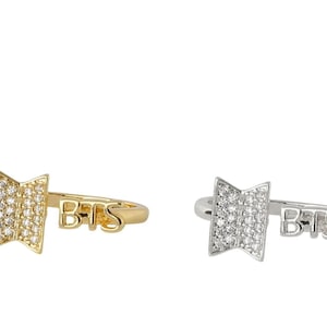 May include: Two rings, one gold-toned and one silver-toned, each featuring a stylized emblem encrusted with small, clear stones. The word "BTS" is inscribed on the band of each ring. The rings are displayed against a white background.