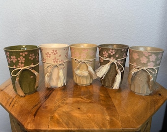 May include: Five ceramic tumblers in varying shades of green, beige, and brown, each adorned with pink floral patterns. The tumblers are tied with a decorative string and tassel. The cups are displayed on a wooden surface.
